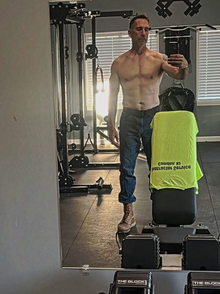 Brian’s later progress photo, standing in his home gym in work jeans and boots